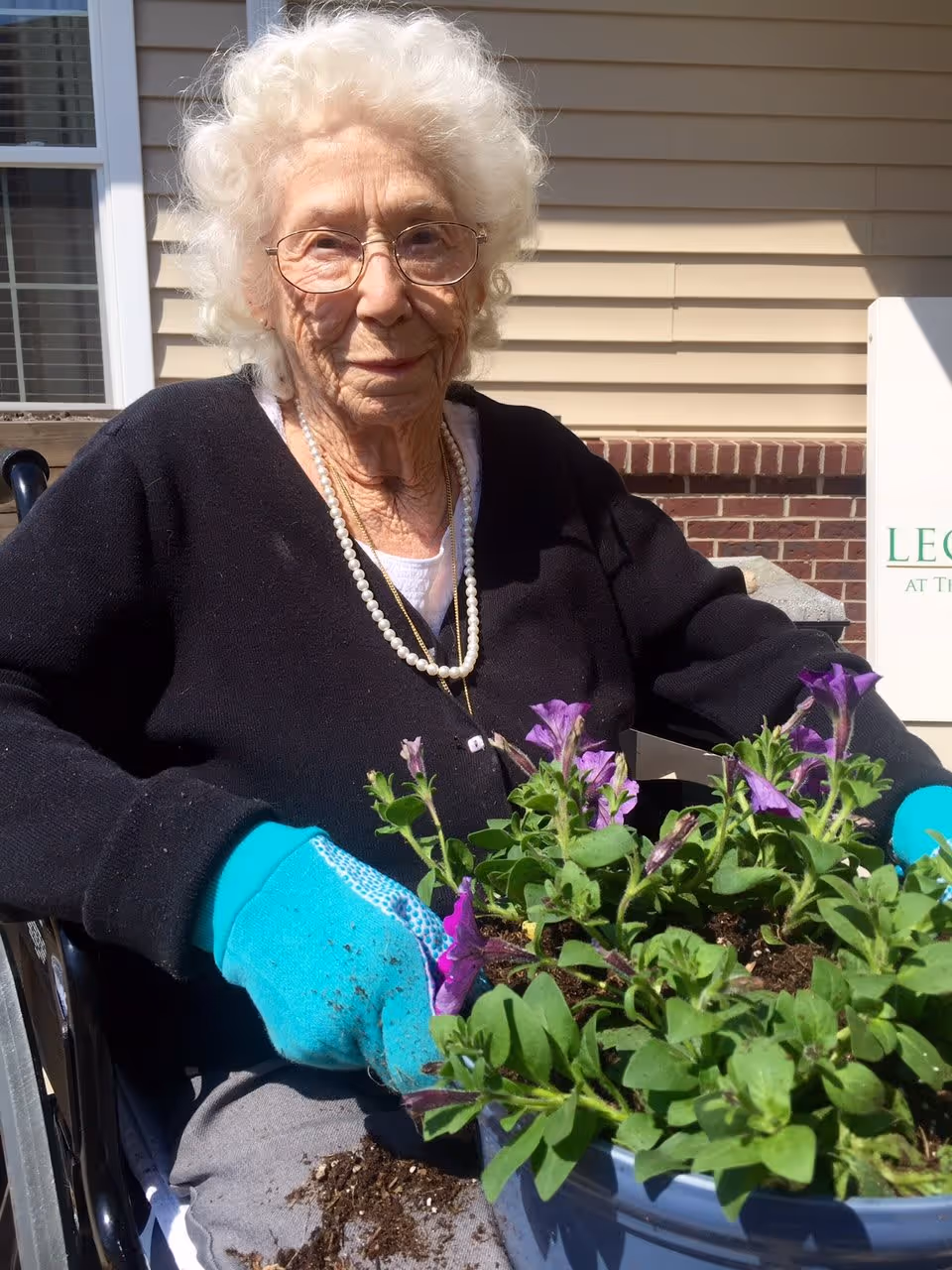 An elderly woman with white curly hair and glasses is sitting in a wheelchair outdoors, wearing a black sweater, a pearl necklace, and bright blue gardening gloves. She is holding a pot filled with green plants and purple flowers, with some soil spilled on her lap. The background shows a beige siding wall with a window and a partial sign.