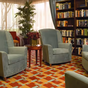 A cozy seating area with four green upholstered armchairs arranged around a small wooden table with a flower arrangement. Behind the chairs is a tall bookshelf filled with books and a large potted plant near a window with sheer curtains allowing natural light to fill the room.