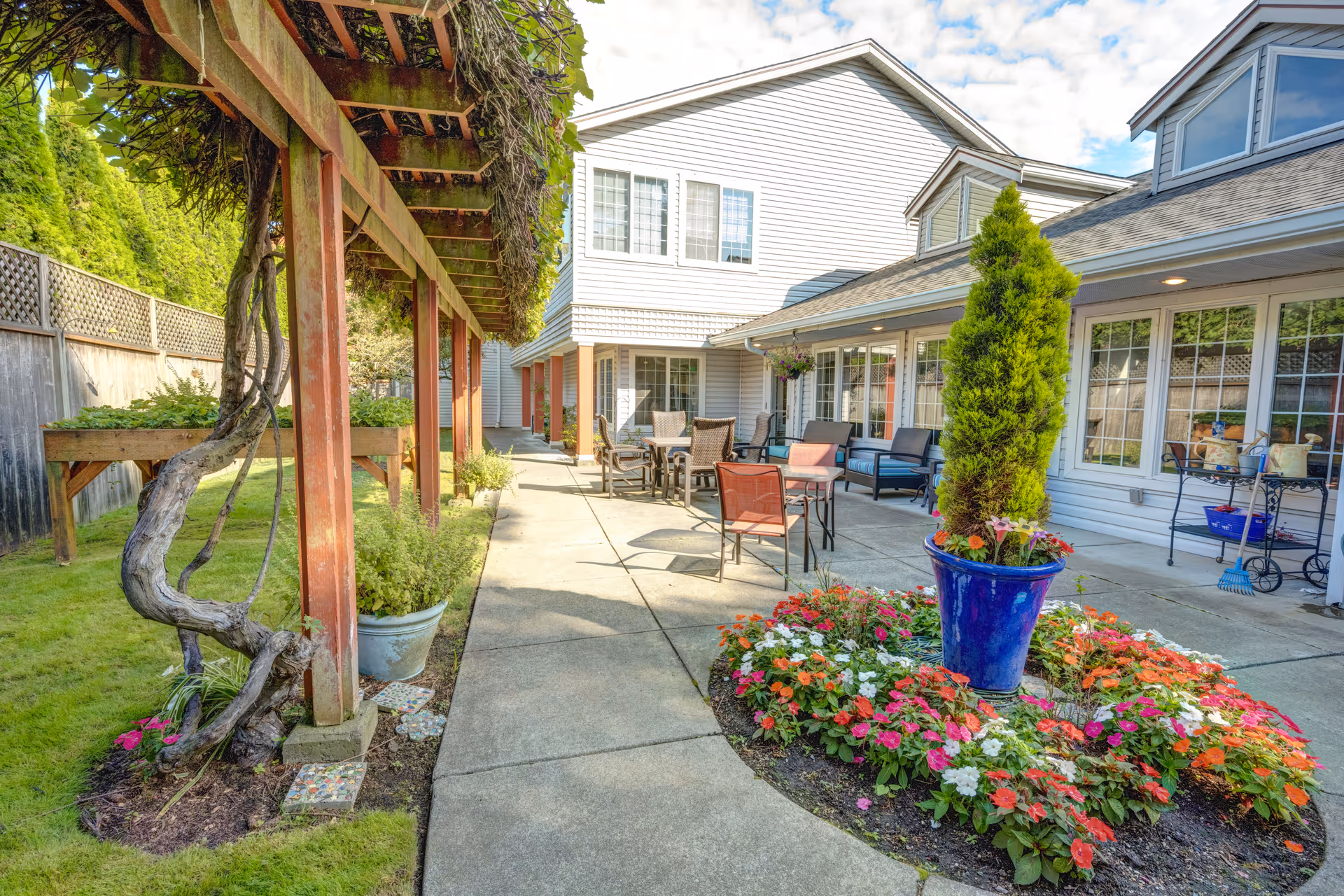 Outdoor patio area at The Terrace at Beverly Lake by Cogir featuring a paved walkway, a pergola with climbing plants, a flower bed with colorful flowers and a tall potted plant, several chairs and tables for seating, and the exterior of a light-colored building with large windows.