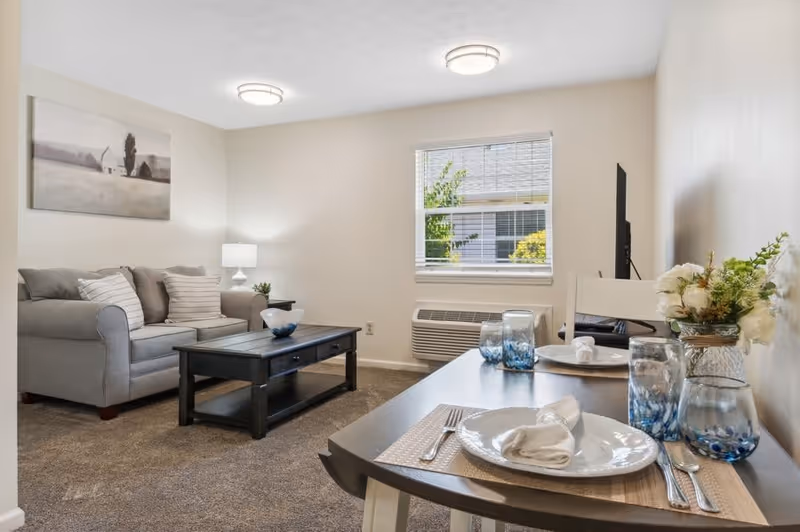 A cozy living room and dining area in a senior living facility. The living room features a gray sofa with striped pillows, a black coffee table, a side table with a lamp, and a wall painting of a barn. The dining area has a table set for two with white plates, napkins, silverware, blue-tinted glasses, and a small floral arrangement. A window with blinds lets in natural light, and an air conditioning unit is installed below the window.