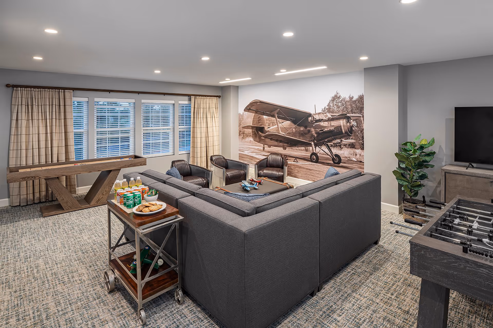 A cozy lounge area with a large gray sectional sofa, three black leather chairs, a coffee table with model airplanes, and a foosball table. There is a large sepia-toned mural of an old airplane on the wall. A wooden shuffleboard table is positioned near windows with beige curtains. A small serving cart with snacks and drinks is next to the sofa. The room has recessed lighting and a carpeted floor.
