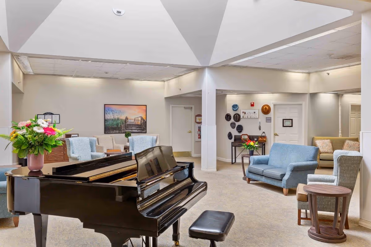 A spacious and well-lit common area in a senior living facility featuring a black grand piano with a matching bench in the foreground. The room has comfortable seating including blue and beige armchairs and sofas, a small wooden side table, and a vase with colorful flowers on the piano. The walls are light-colored with a framed picture of a boardwalk at sunset and a collection of hats hanging near a door. The ceiling has recessed lighting and a skylight.
