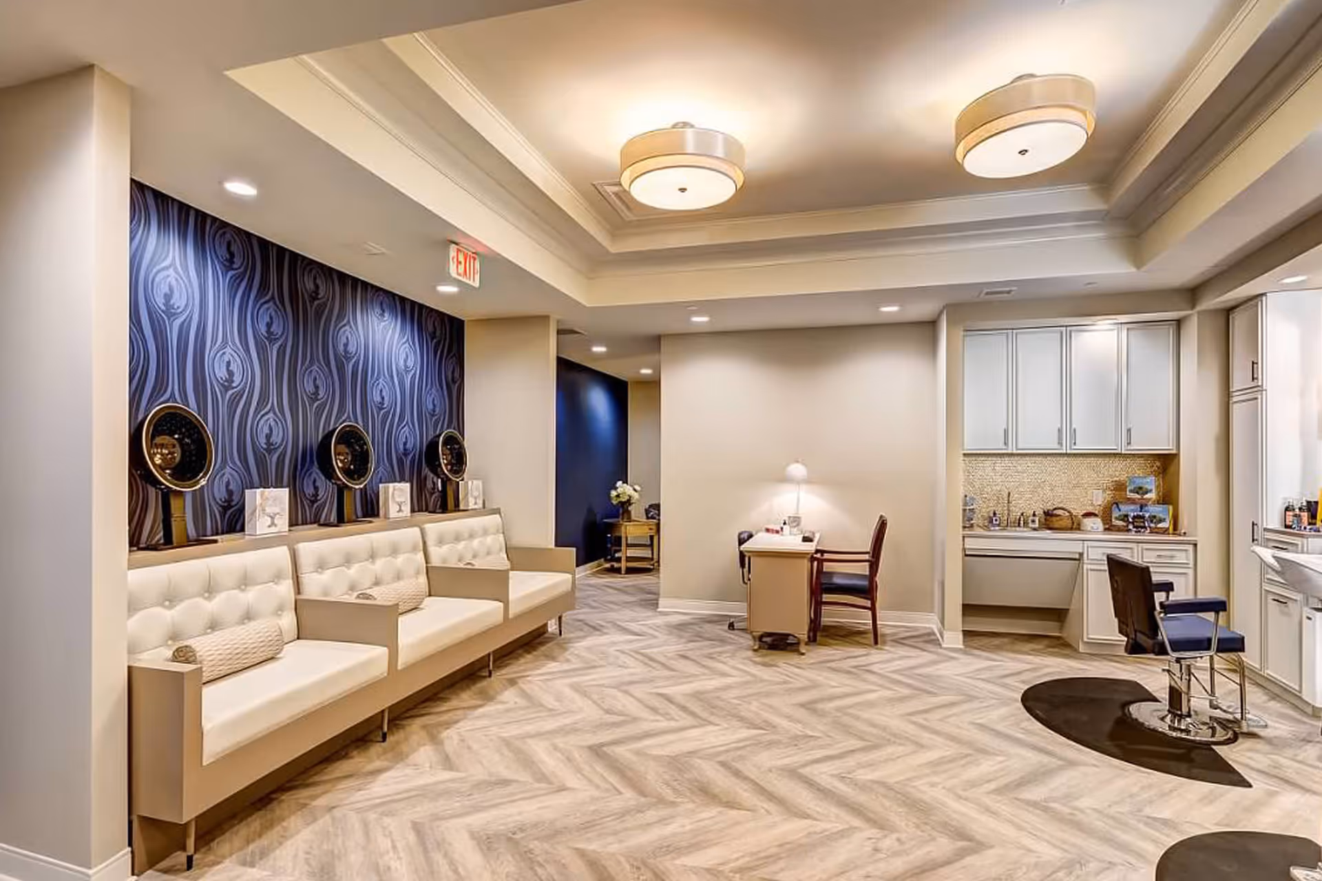 Interior view of a stylish salon area in a senior living facility with three vintage hair dryers mounted on a wall with dark patterned wallpaper, cream-colored cushioned bench seating beneath them, a small desk with a chair and lamp, and a hair styling station with a chair and cabinetry in the background.