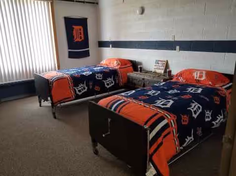 Two twin beds with matching Detroit Tigers–themed bedding in a simple senior living bedroom with a small dresser and vertical blinds.