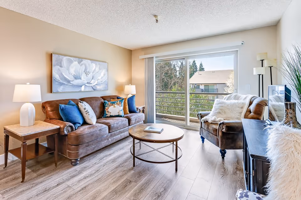 A bright living room with a brown leather sofa and armchair, both adorned with decorative pillows and a white throw. There is a round wooden coffee table in the center, two side tables with lamps, a large floral painting on the wall, and a sliding glass door leading to a balcony with a view of trees and neighboring buildings.