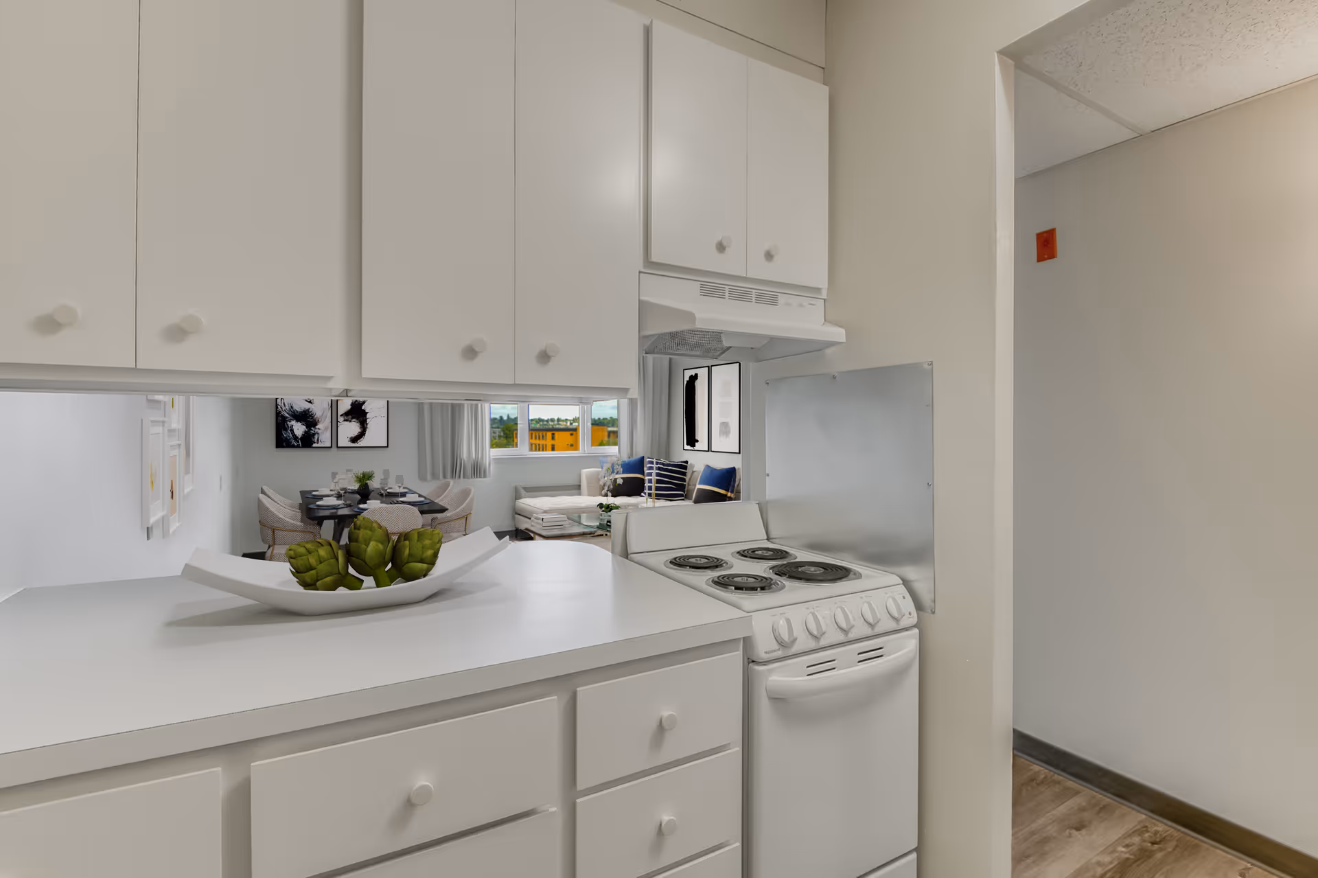 A small kitchen area with white cabinets and a white electric stove. A white countertop holds a decorative white dish with green artichokes. Through a pass-through window, a dining area with a round table and chairs, and a living room with a couch and cushions are visible. The walls are light-colored, and there is a window showing an outdoor view.