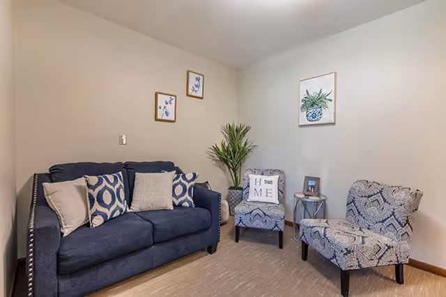 A cozy living room with a dark blue sofa adorned with patterned and plain cushions, two patterned armchairs, a small round side table with a framed photo, a tall green plant in the corner, and three framed botanical prints on the walls.