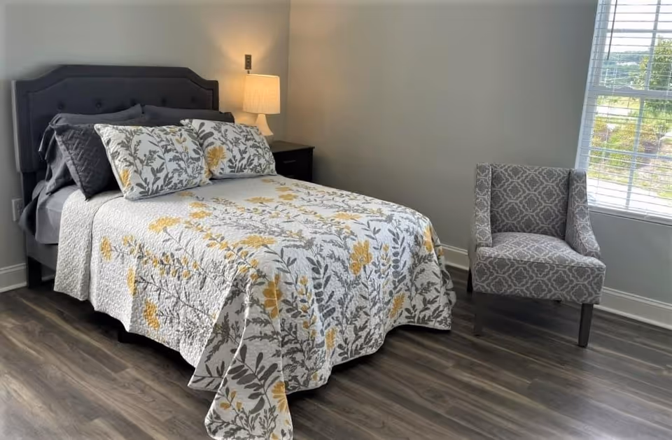 A neatly made bed with a floral patterned quilt and matching pillows in shades of yellow and gray. Next to the bed is a dark nightstand with a lit table lamp. A gray patterned armchair is positioned near a window with white blinds, allowing natural light to brighten the room. The floor is covered with wood-style flooring.