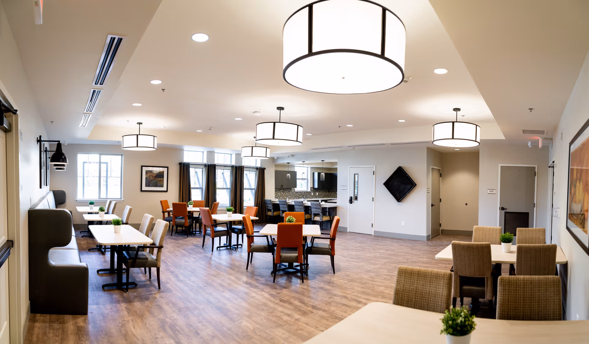 A spacious, well-lit dining room with multiple tables and chairs arranged neatly. The room features large windows with dark curtains, modern ceiling lights, and a wooden floor. Small green plants are placed on the tables, and there is a kitchen area visible in the background.