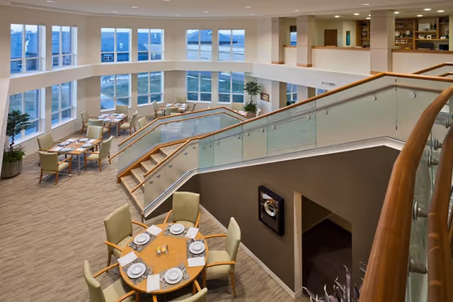 Spacious and modern dining area in a senior living facility with multiple round tables set with plates, napkins, and utensils. The room features large windows allowing natural light, beige walls, and a staircase with glass and wood railing leading to an upper level.