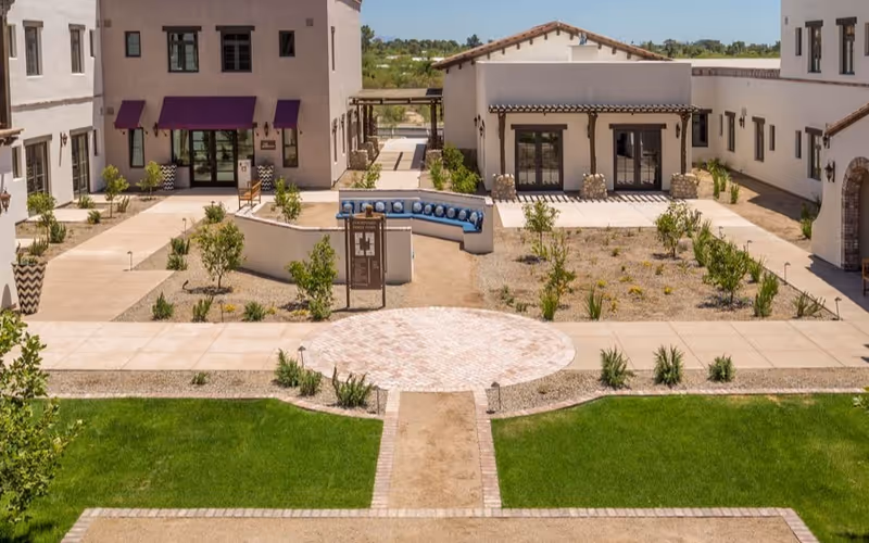 Central courtyard with paved paths, lawns, planted beds, a curved blue-seated bench, and surrounding stucco buildings with awnings.