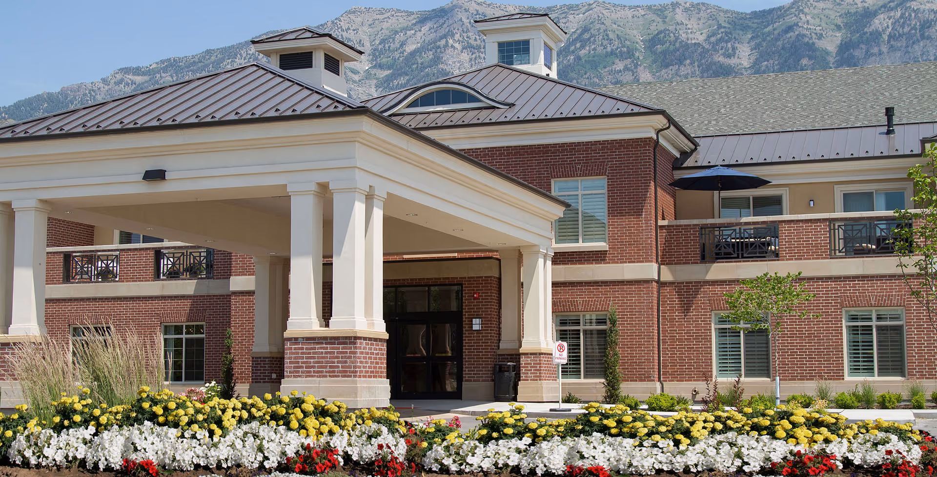 Front exterior view of Legacy Village of Provo, showing a brick building with a covered entrance, landscaped flower beds with yellow, white, and red flowers, and mountains in the background.