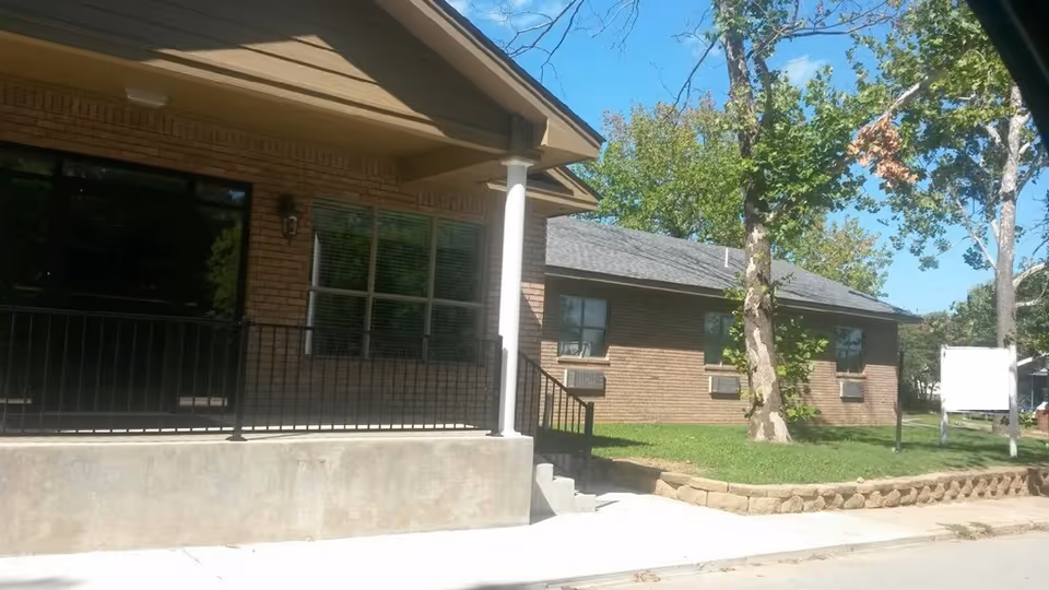 Exterior view of a brick building with a covered porch supported by white columns, black metal railing, and steps leading to a sidewalk. There are trees and a grassy area with a retaining wall in front of the building under a clear blue sky.