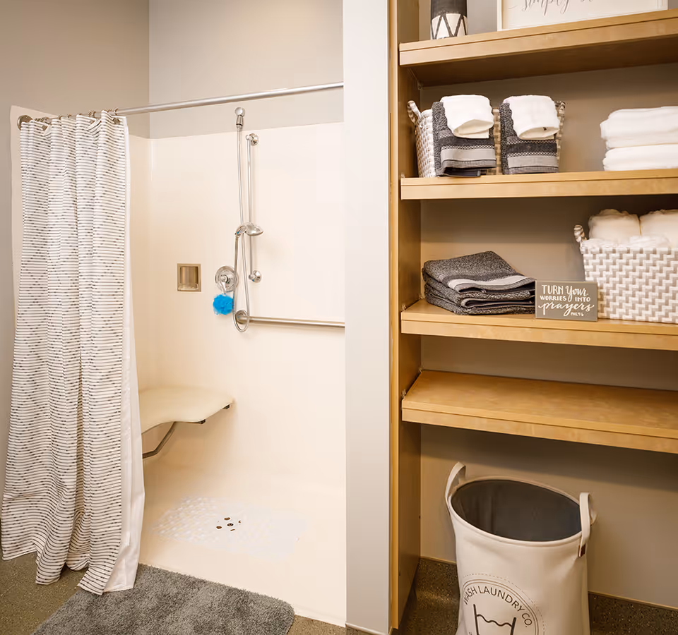 A bathroom shower area with a white shower curtain featuring thin horizontal stripes. The shower has a built-in fold-down seat, a handheld showerhead, and a grab bar. Next to the shower is a wooden shelving unit with neatly folded towels, baskets, and a small sign that reads 'TURN your WORRIES INTO prayers'. A laundry hamper is placed on the floor beneath the shelves.