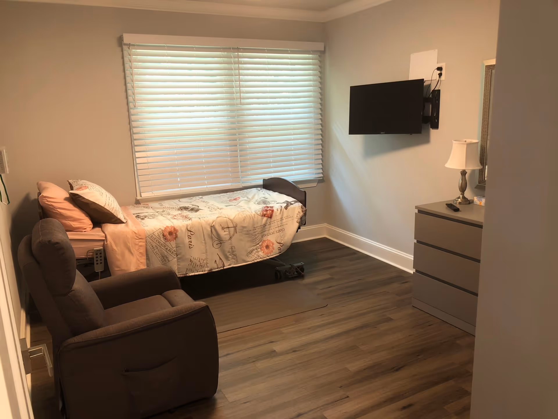 A senior living bedroom with a single bed covered in a floral and script patterned bedspread, a brown recliner chair, a window with closed blinds, a wall-mounted flat screen TV, and a gray dresser with a lamp and remote control on top.