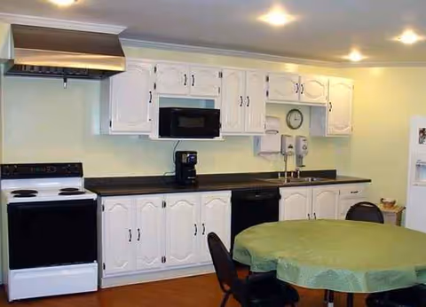 White kitchen cabinets line the wall with appliances and a round table in foreground.