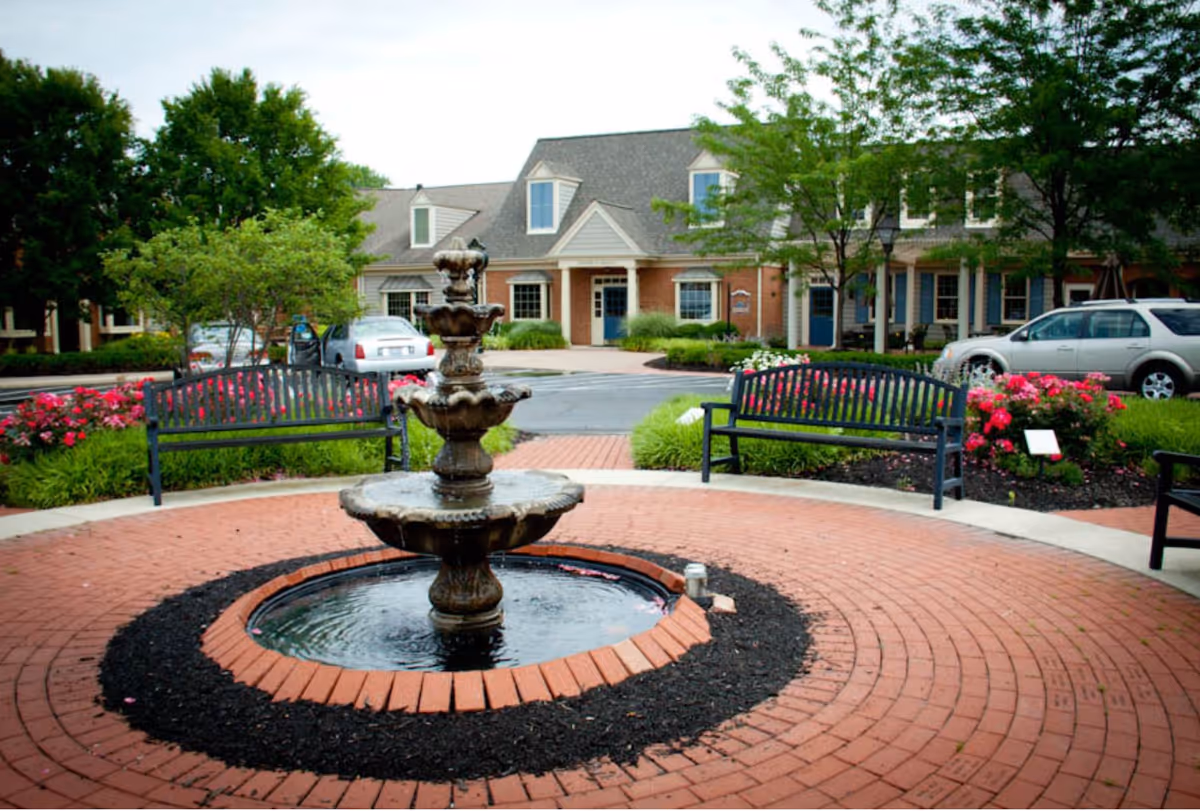 Outdoor courtyard area with a multi-tiered water fountain in the center, surrounded by a circular brick pathway. There are black metal benches and flower beds with pink flowers and green shrubs. In the background, there is a building with a gray roof, dormer windows, and a brick facade. Several cars are parked near the building.