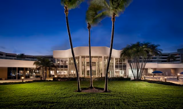 Front exterior of Harbour's Edge building at dusk with illuminated glass entrance, palm trees, and a manicured lawn.