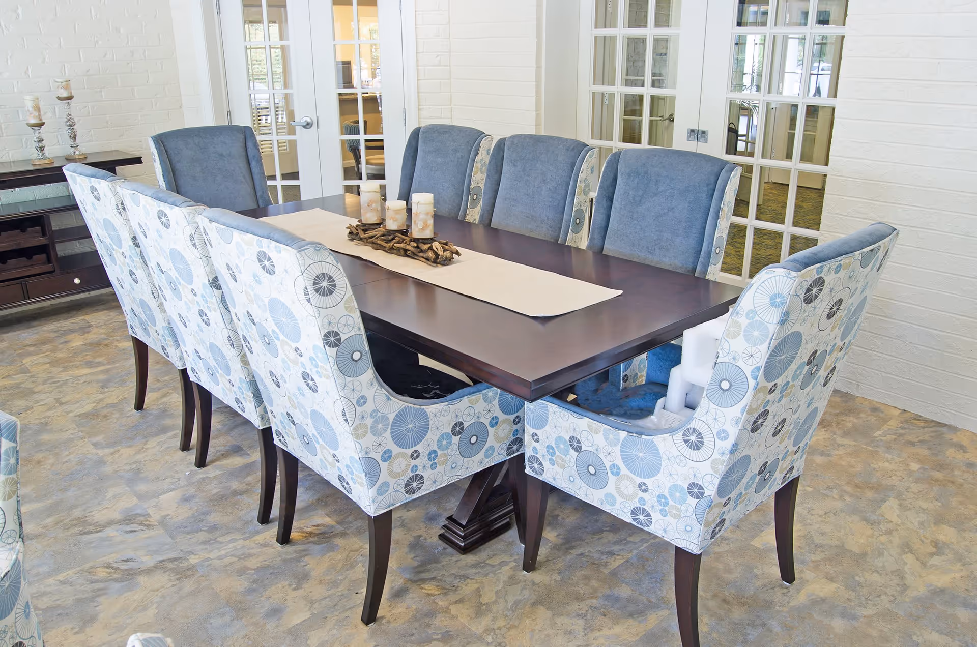 A dining room with a dark wooden rectangular table surrounded by eight upholstered chairs. The chairs have a patterned fabric with blue, gray, and beige circular designs on the outside and solid blue fabric on the inside. The table has a beige runner and a centerpiece with candles and decorative driftwood. The room has a light-colored floor and white brick walls with glass-paneled double doors in the background.