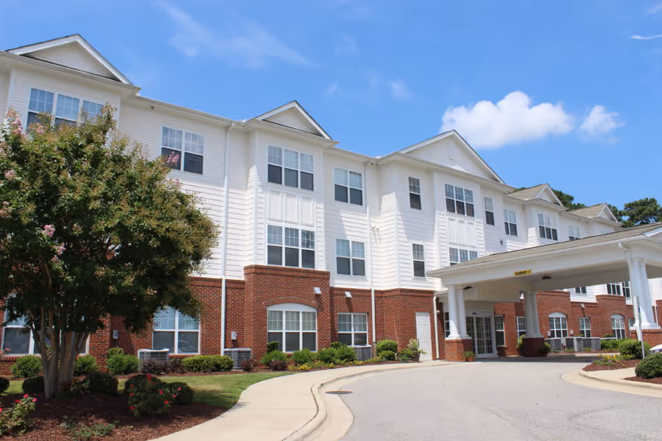 Front view of a three-story white and red-brick senior living building with a covered entrance, landscaped grounds, and a clear blue sky.