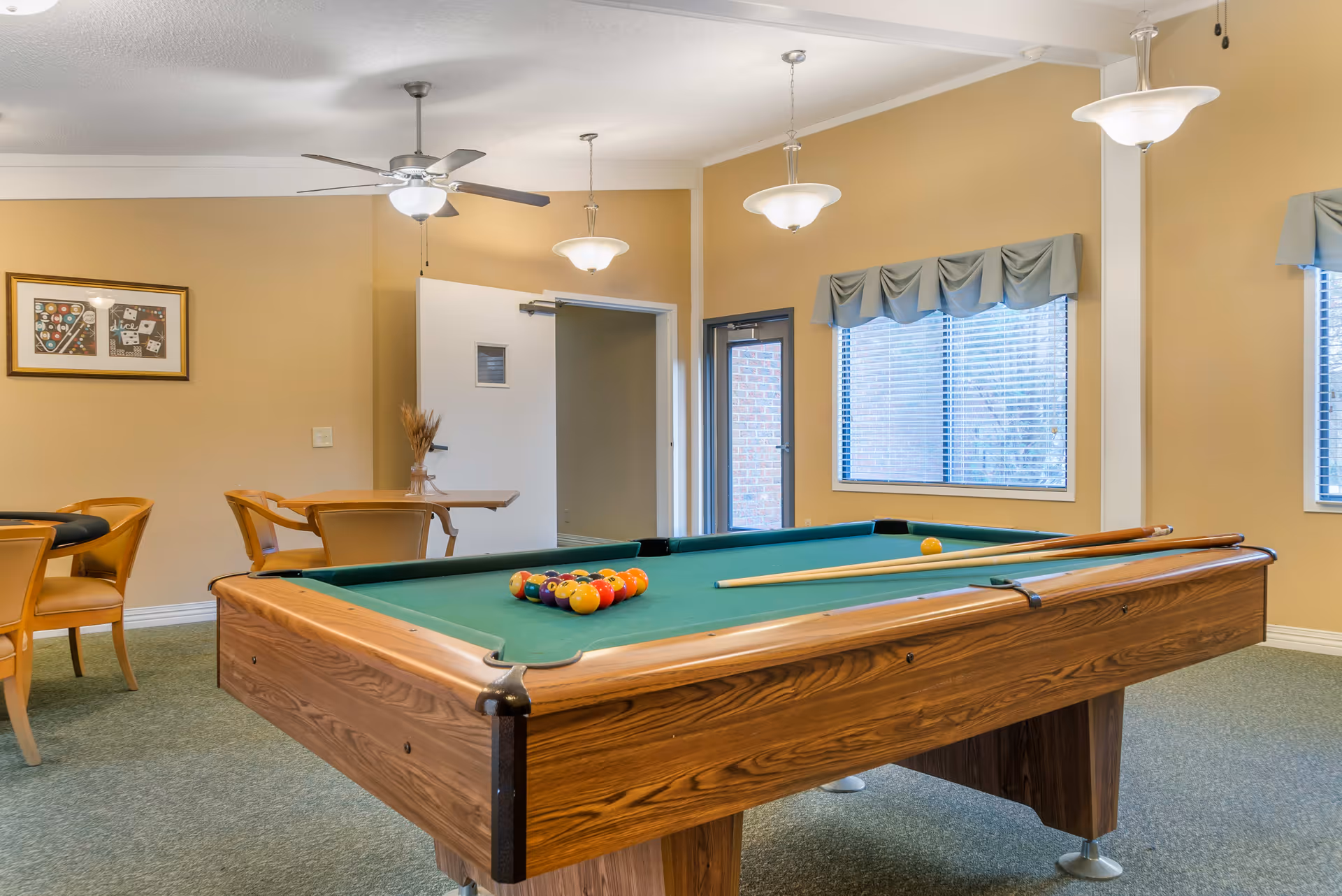 A recreational room with a pool table in the center, set up with billiard balls and cues. The room has beige walls, green carpet, ceiling fans with lights, and large windows with blue valances. There are also tables and chairs along the walls, and a framed picture on one wall.