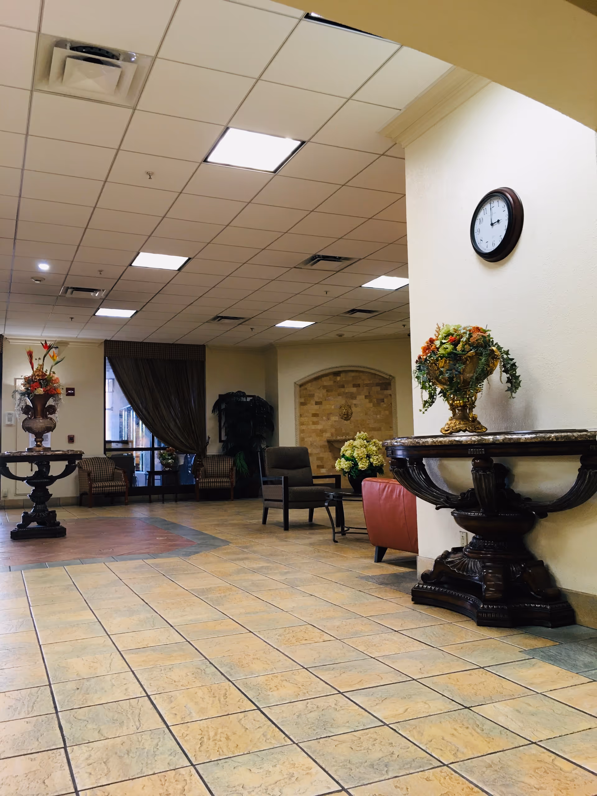 Interior view of a nursing and rehabilitation center lobby with tiled floor, decorative tables with flower arrangements, chairs, a clock on the wall, and large windows with curtains.