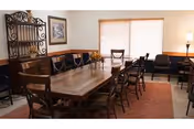 Communal dining room with a long wooden table surrounded by chairs, a sideboard and wall decor under a window with blinds.