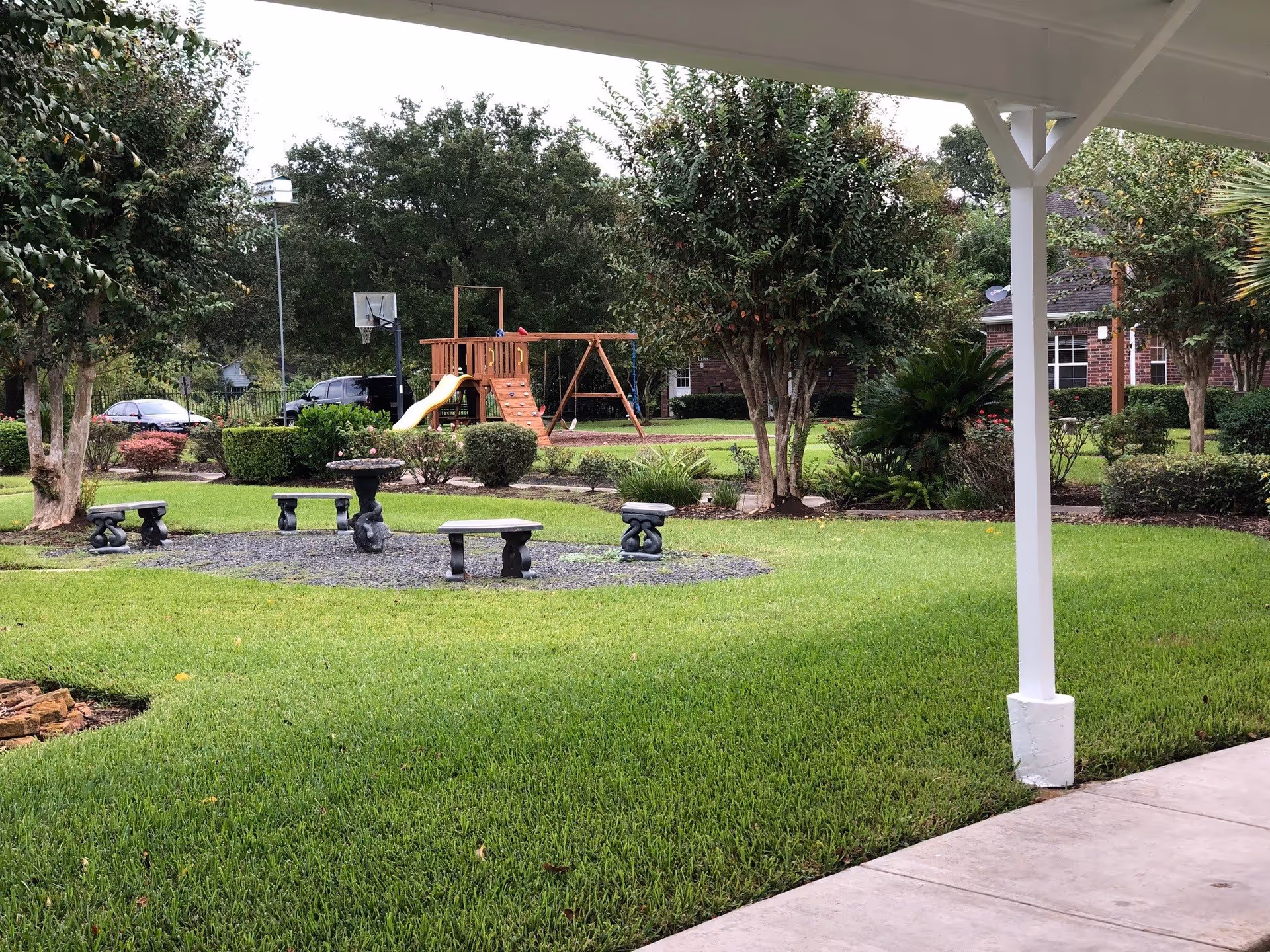 Courtyard with green lawn, stone benches and a birdbath, a wooden playset with slide, and trees seen from a covered walkway.