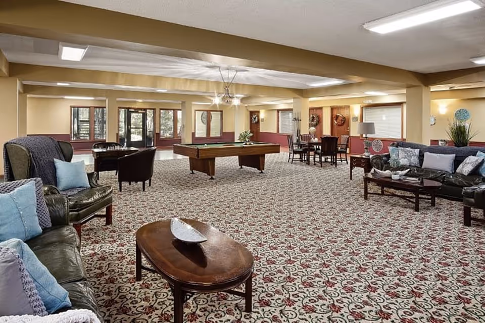 Spacious carpeted common lounge with sofas, chairs, a pool table and dining tables in a senior living facility.