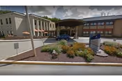 Covered main entrance of a healthcare facility with a driveway, parked car, landscaping and a freestanding sign.