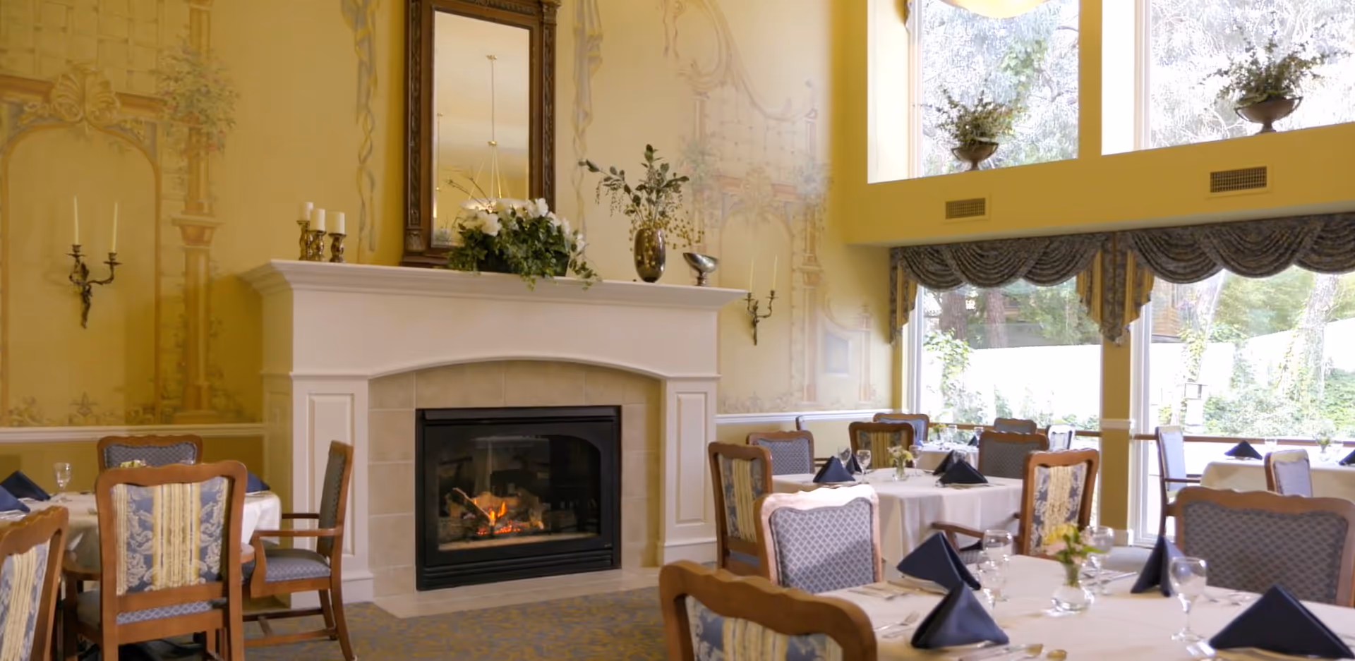 A cozy dining room with several round tables covered in white tablecloths, each set with glassware, silverware, and navy blue folded napkins. The room features a lit fireplace with a white mantel decorated with flowers and a mirror above it. The walls are painted yellow with intricate designs, and large windows with valances let in natural light, showing greenery outside.