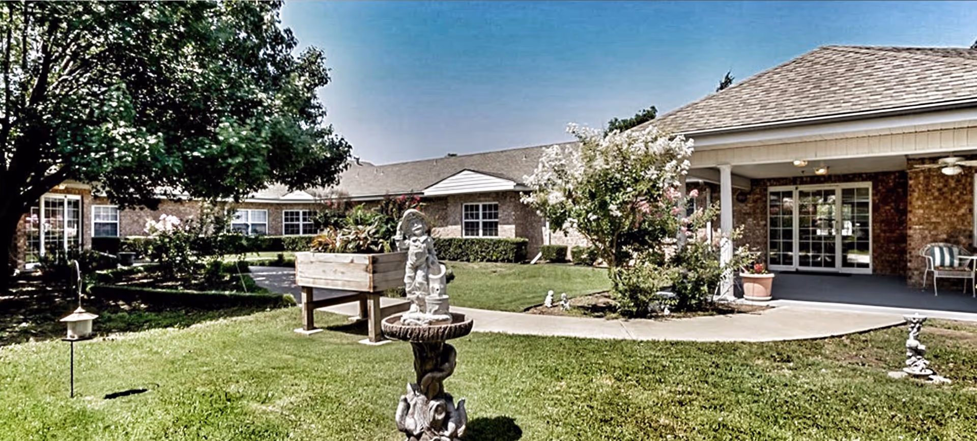 Outdoor courtyard area of Countryside Senior Living of Greenville featuring a well-maintained lawn, a decorative statue fountain, a raised wooden planter box with plants, trees, and shrubs. The building has brick walls, multiple windows, and a covered patio with chairs and a ceiling fan.