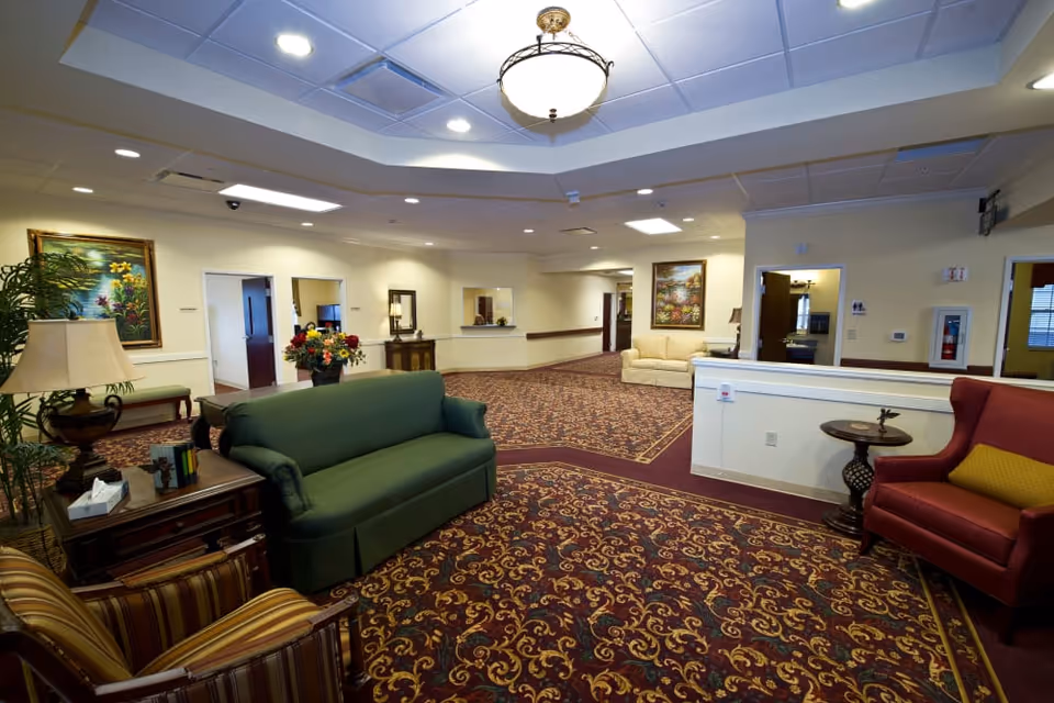 Spacious senior living facility lobby with upholstered seating, patterned carpet, paintings, and a reception desk.