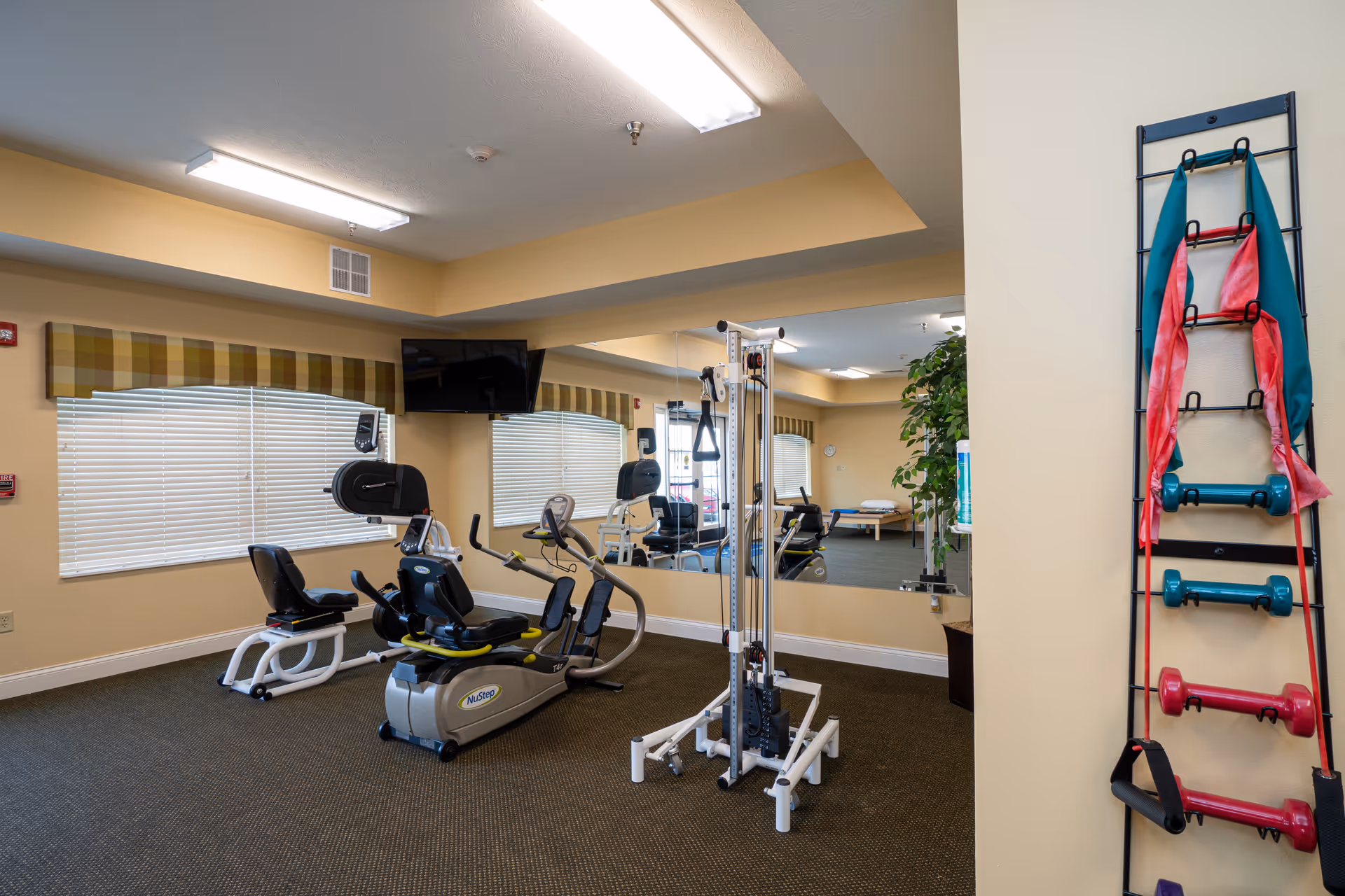 A small fitness room with exercise equipment including a recumbent bike, a multi-function weight machine, and a rack holding resistance bands and dumbbells. The room has beige walls, a large mirror on one wall, windows with blinds and plaid valances, and fluorescent ceiling lights.