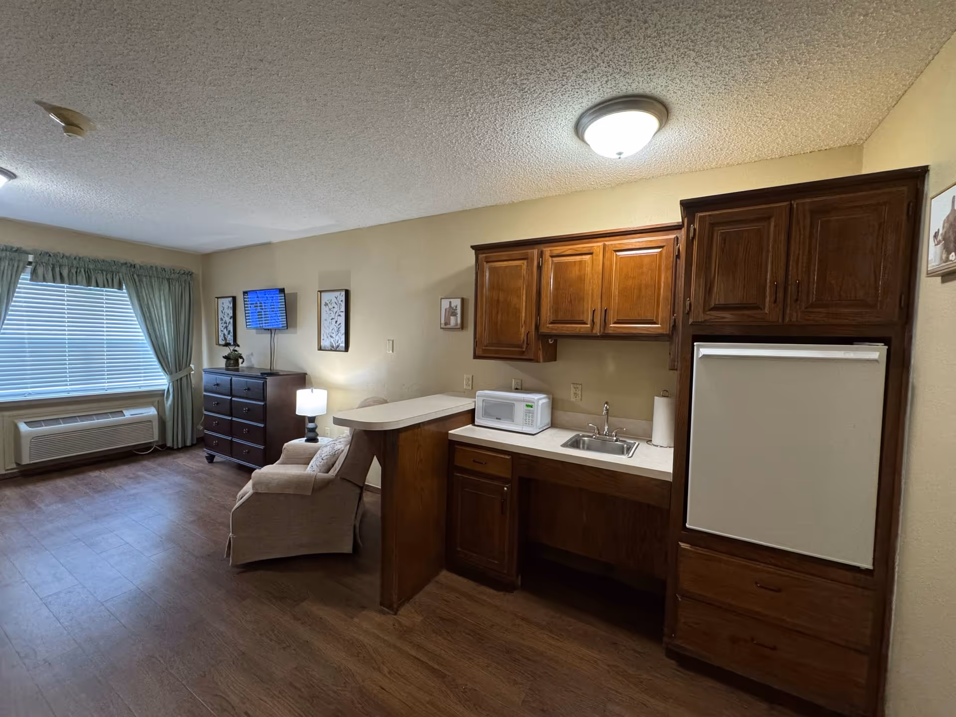 Interior view of a senior living facility room at Preston Place featuring a small kitchenette with wooden cabinets, a microwave, a sink, and a refrigerator. Adjacent to the kitchenette is a cozy sitting area with an armchair, a dresser with a lamp, and a wall-mounted TV. A large window with green curtains and an air conditioning unit below it is visible on the left side of the room.