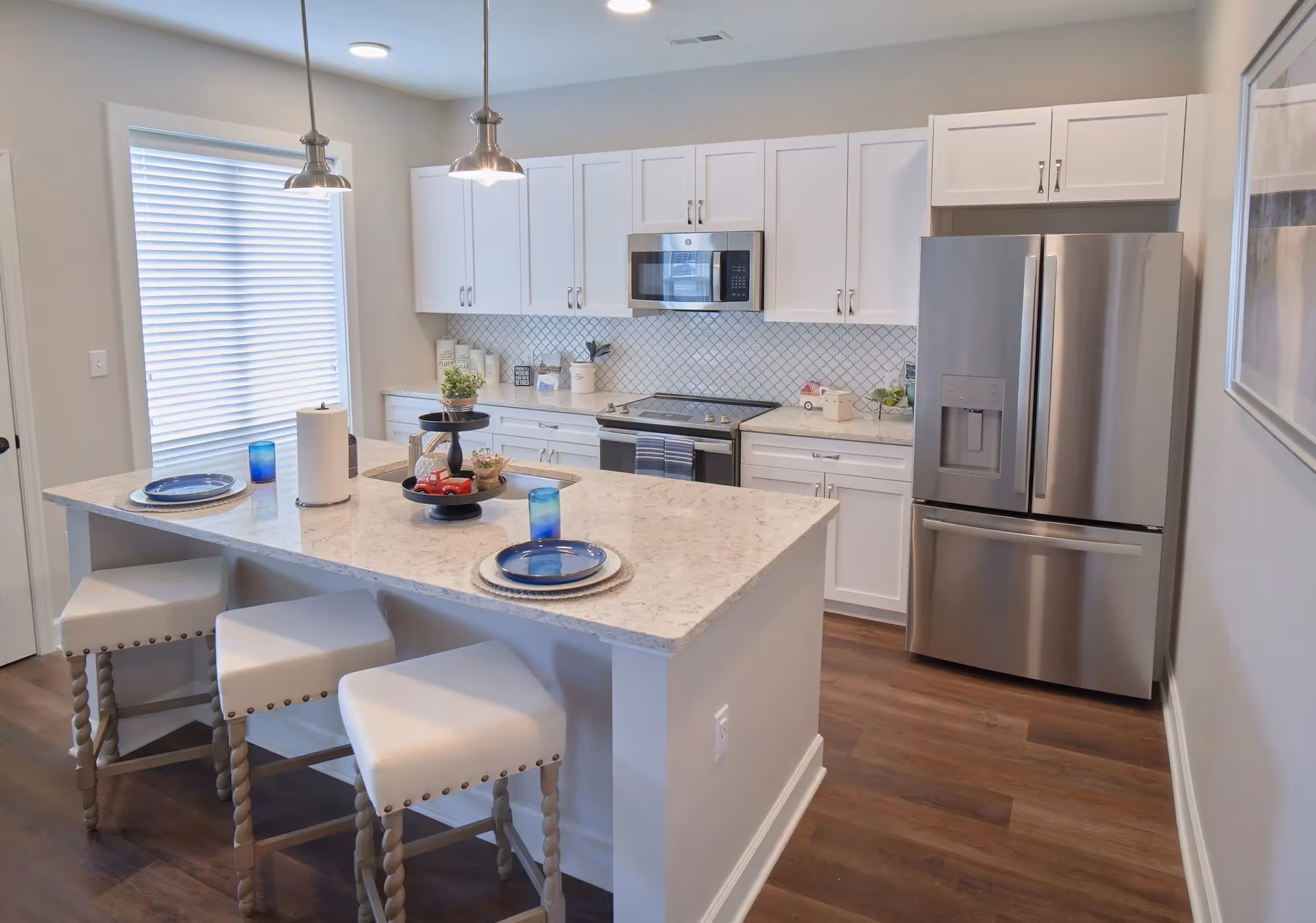 Modern kitchen with white cabinets, a stainless steel refrigerator, microwave, and stove. There is a kitchen island with a marble countertop, three cushioned stools, two blue plates with glasses, and pendant lights hanging above. The floor is wooden and there is a window with blinds on the left side.