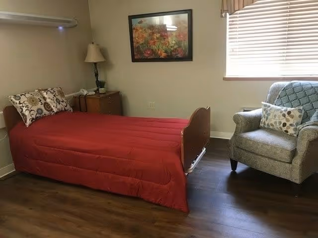 Small furnished bedroom with a single bed covered in a red comforter, a nightstand and lamp, a framed floral painting, and an armchair by a window.