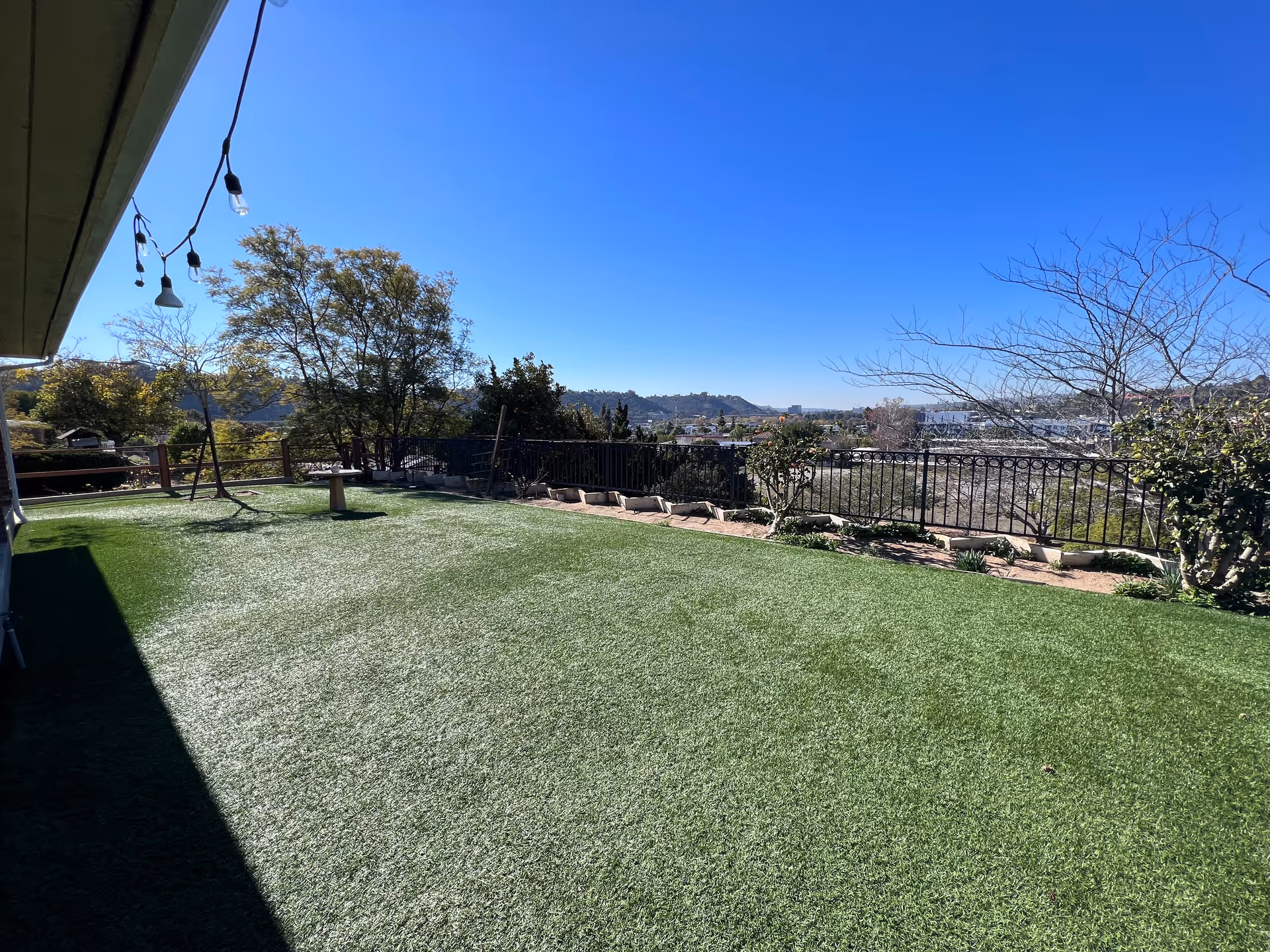 Sunlit backyard with artificial turf, string lights, trees and a decorative railing overlooking distant hills and the city.