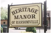 A large rectangular sign with a cream background and black border that reads 'HERITAGE MANOR OF RANSOMVILLE' mounted on two black posts outdoors with trees and buildings in the background.