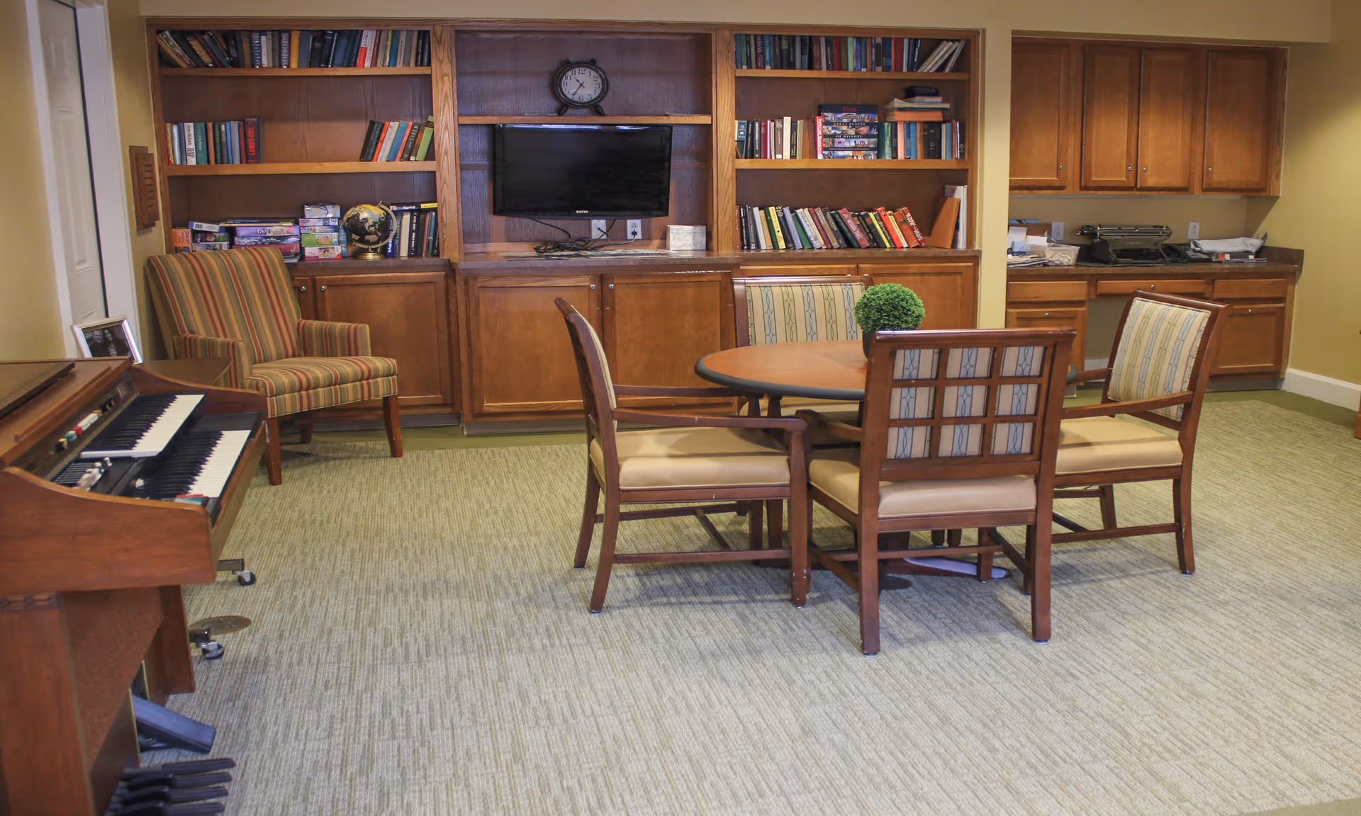 A communal sitting room with a round table and chairs, bookshelves and TV on the back wall, a striped armchair and a piano to the side.