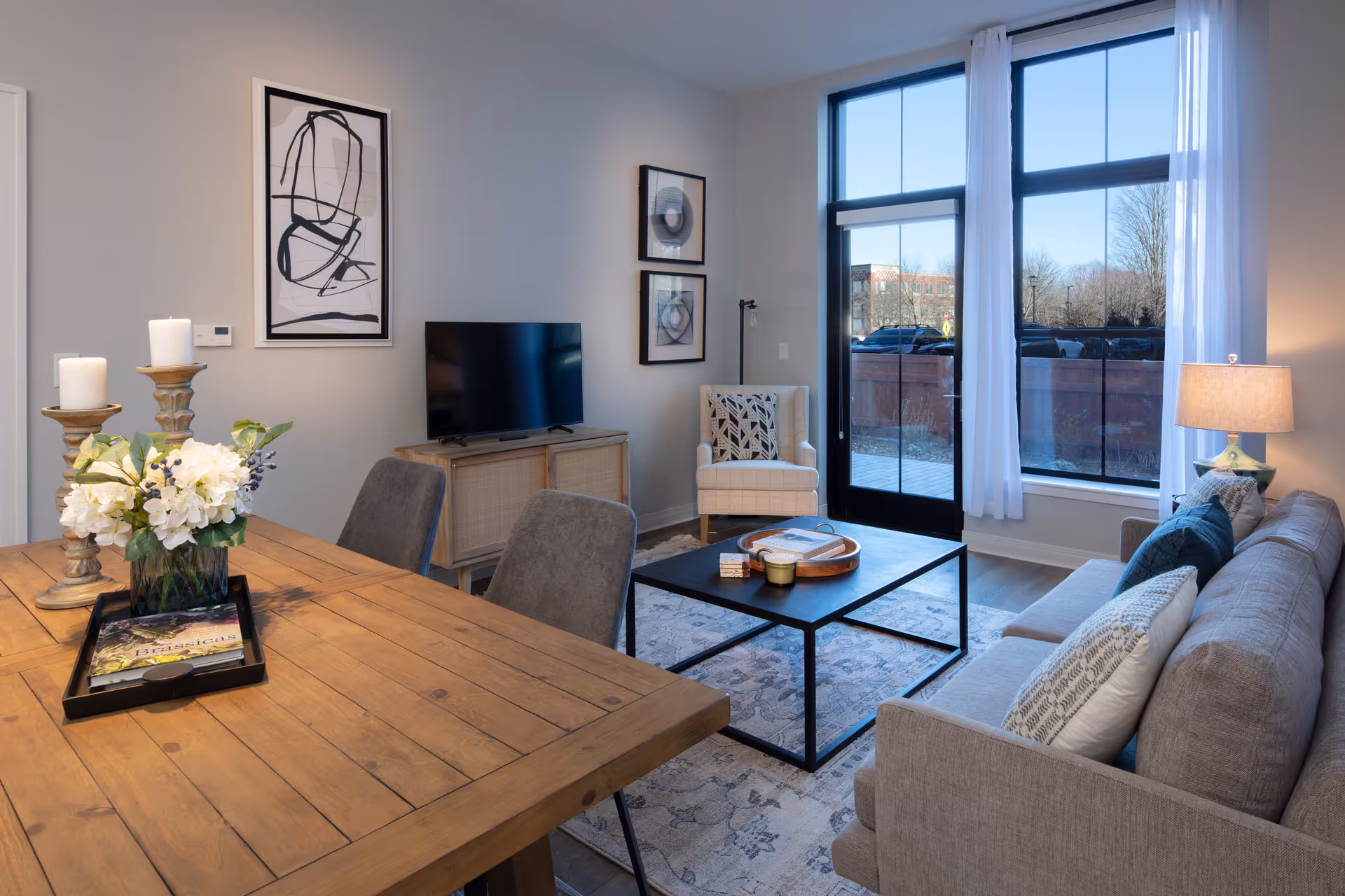 A modern living room and dining area with a wooden dining table and gray chairs in the foreground. The living room features a gray sofa with pillows, a black metal coffee table on a patterned rug, a beige armchair, a TV on a wooden stand, and large windows with white curtains letting in natural light.