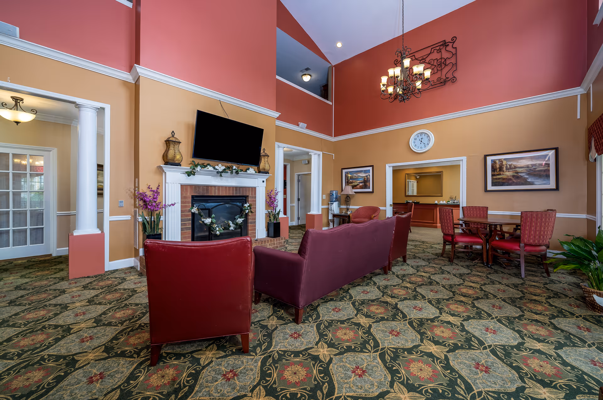 A senior living facility common area with a patterned carpet, red and beige walls, and a high ceiling. The room features a fireplace with a TV mounted above it, flanked by decorative lanterns and floral arrangements. There are red upholstered chairs and a sofa arranged around the fireplace, a round wooden table with four red chairs, framed landscape paintings on the walls, a clock, a chandelier, and a water dispenser on a small table.
