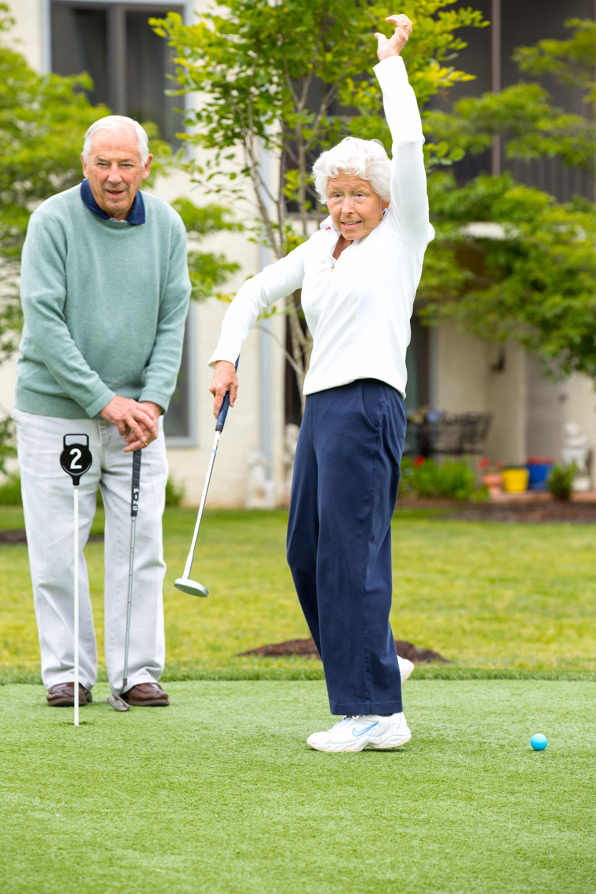 An elderly woman is playing golf on a putting green, raising one arm in celebration or stretch, while an elderly man stands nearby holding a golf club and watching her. They are outdoors with greenery and a building in the background.