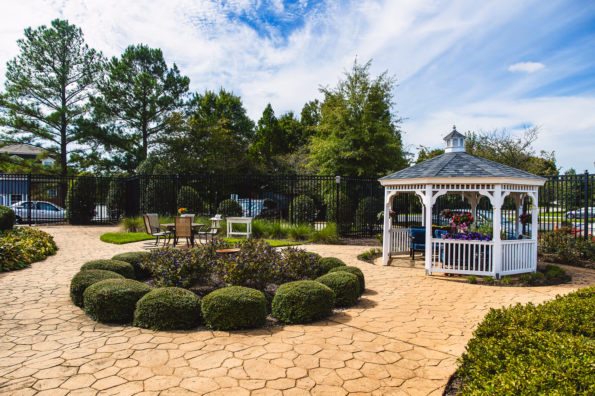 Outdoor garden area with a circular flower bed surrounded by neatly trimmed bushes, a white gazebo with hanging flower pots and seating inside, a small table with chairs, and a black metal fence enclosing the space. Trees and parked cars are visible in the background under a partly cloudy blue sky.