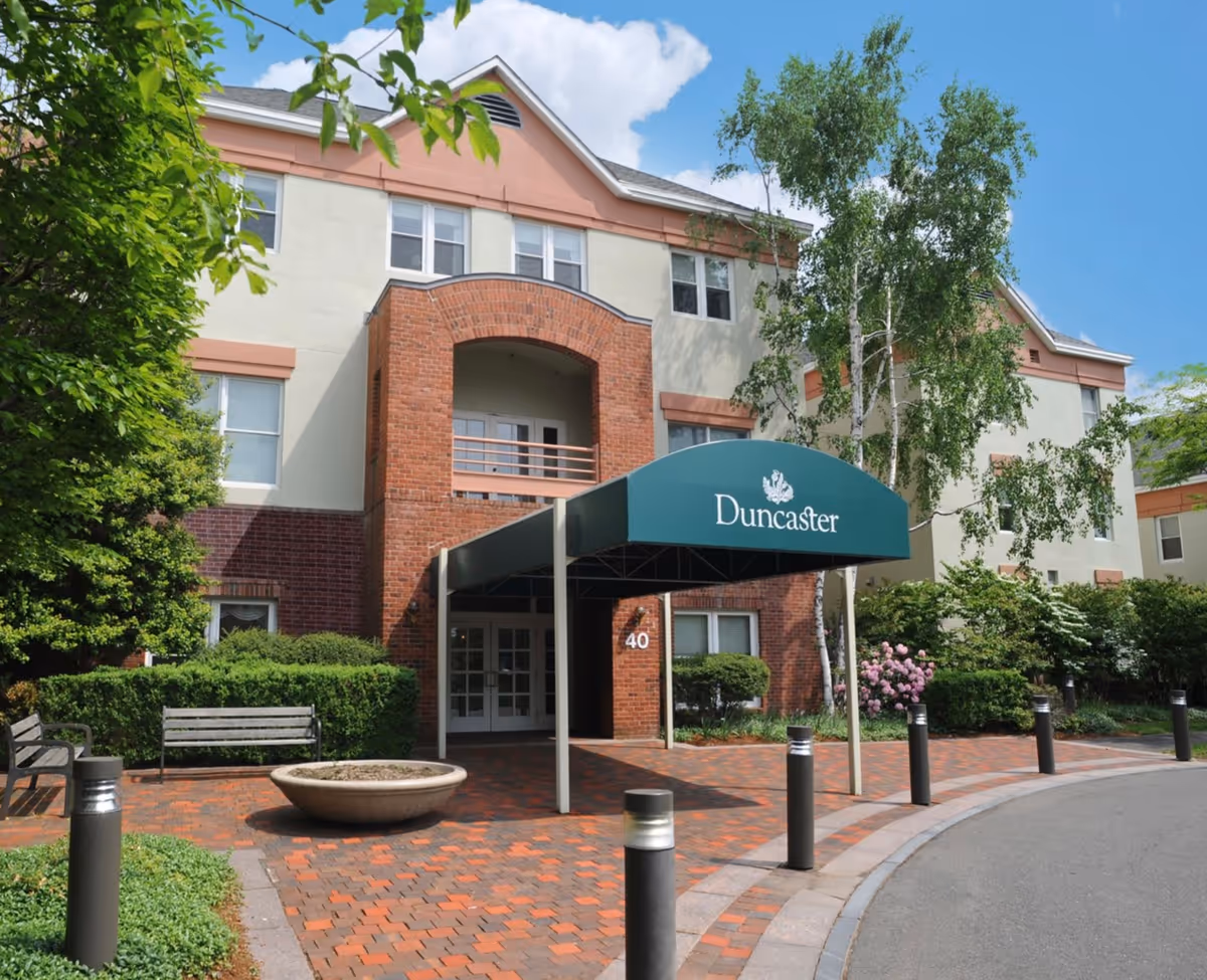 Exterior view of a multi-story senior living facility named Duncaster with a brick and cream facade, a green awning over the entrance, benches, trees, and a clear blue sky.