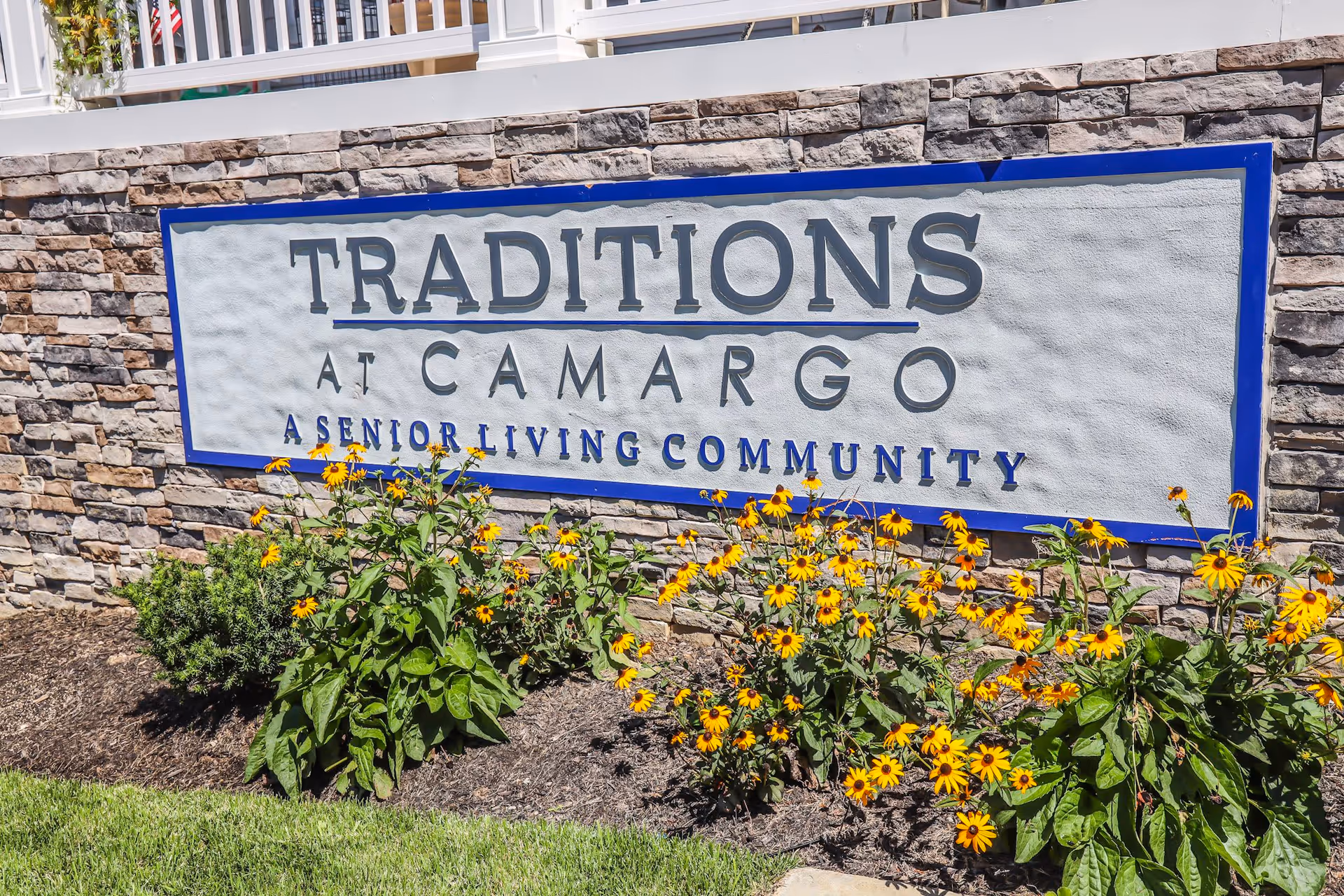 Stone wall with a large sign that reads 'Traditions at Camargo A Senior Living Community' surrounded by yellow flowers and green plants in front of the wall.