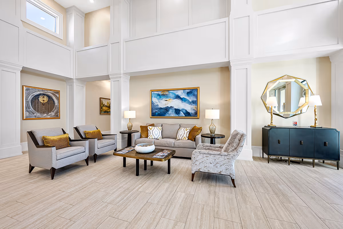 A bright and spacious living room area with high ceilings and large white columns. The room features a beige sofa with decorative pillows, two gray armchairs with yellow cushions, and a patterned armchair. A wooden coffee table with a decorative bowl and magazines sits in the center. On the walls are framed paintings, and a large geometric mirror hangs above a dark blue sideboard with two lamps. The floor is covered with light-colored tiles.