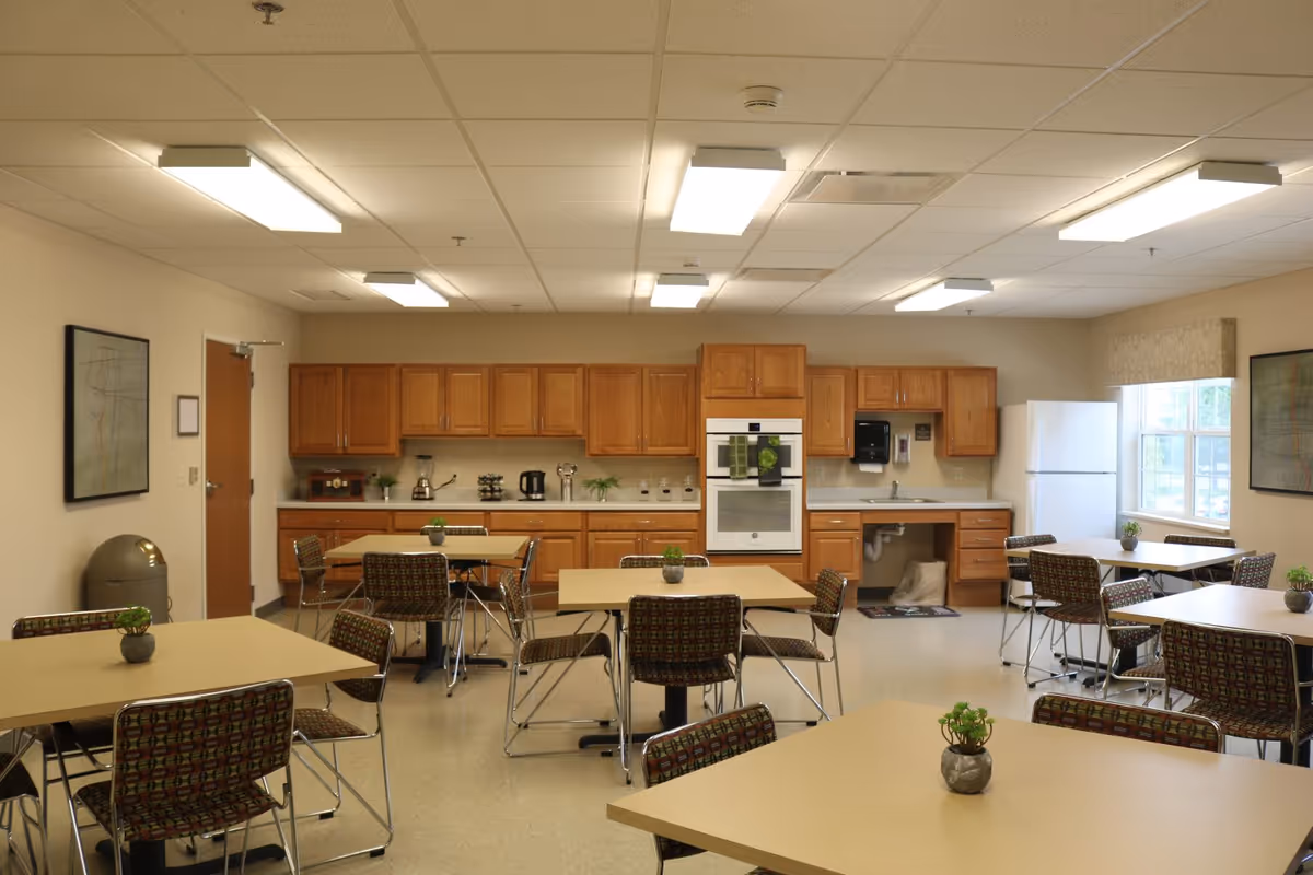A well-lit communal kitchen and dining area with multiple square tables and patterned chairs. The kitchen features wooden cabinets, a white refrigerator, a built-in oven, a sink, and various small appliances on the countertop. Small potted plants decorate the tables, and framed artwork hangs on the walls.