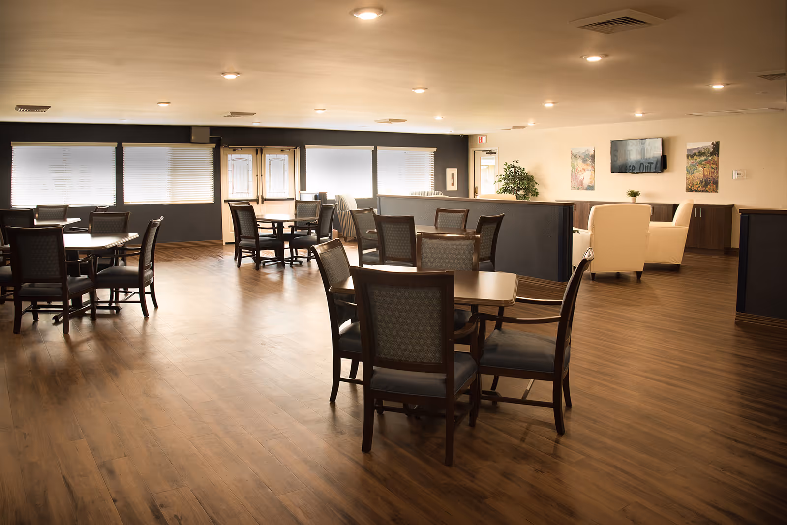 Spacious communal dining and lounge area with wooden floors, multiple tables and chairs, and a seating area with armchairs and a wall-mounted TV.