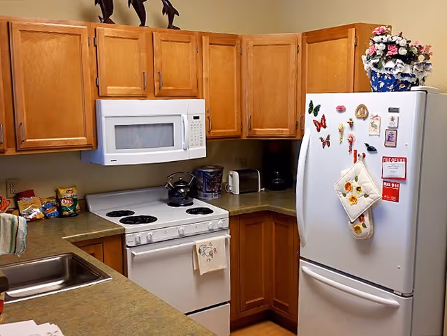 Small kitchen with wooden cabinets, a white stove and microwave, a refrigerator with magnets, and countertops with appliances and snacks.